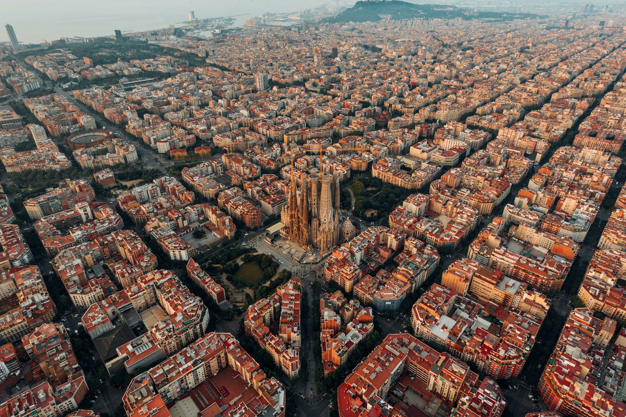 Vista aérea de la Sagrada Familia elevándose sobre Barcelona, con la cálida luz dorada del amanecer proyectando largas sombras sobre la trama urbana y resaltando los tejados circundantes. 