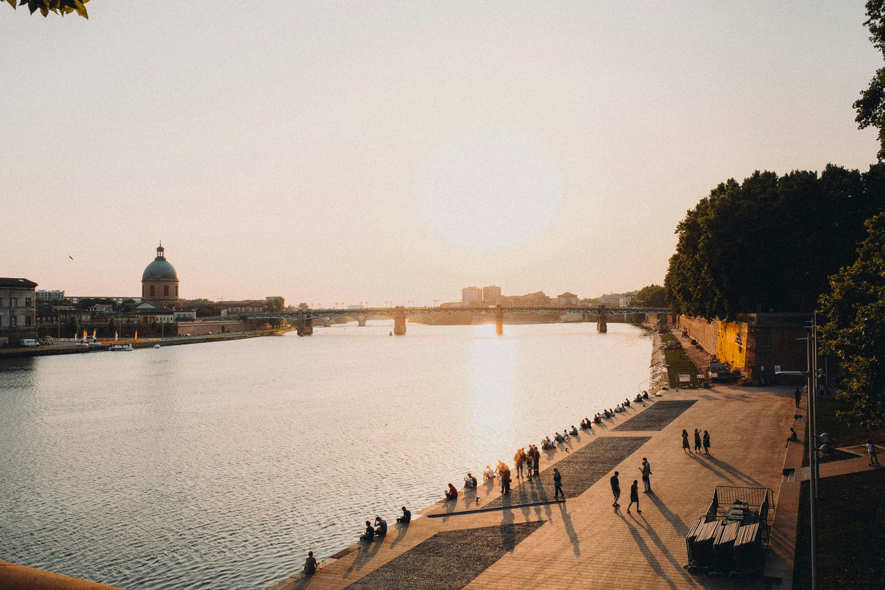 La gente pasea y se relaja a lo largo del río Garona en Toulouse al atardecer, con la cúpula del hospital de La Grave visible al otro lado del agua.