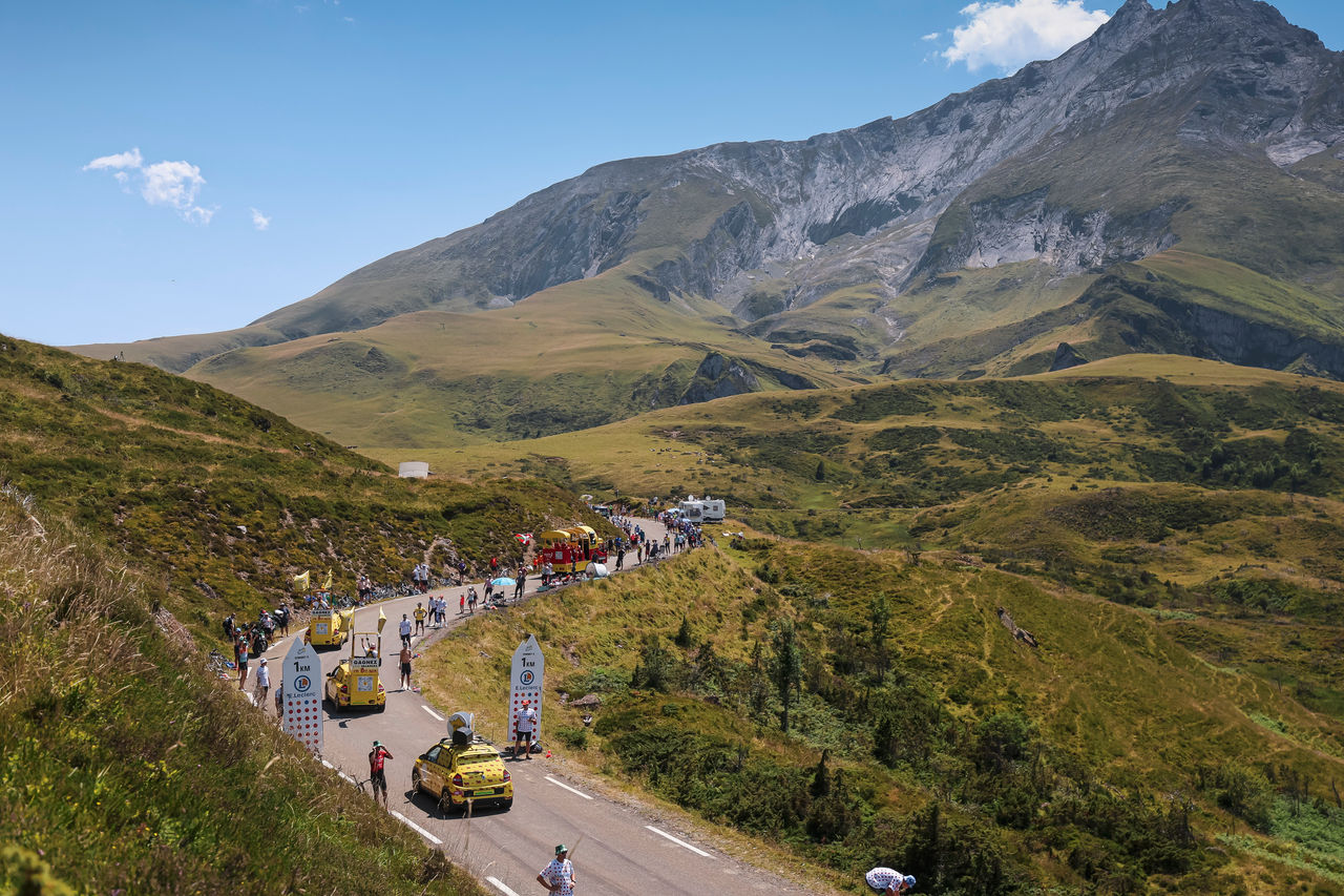 Una carretera de montaña serpenteante, flanqueada por espectadores y vehículos de apoyo, con colinas verdes y un imponente pico rocoso bajo un cielo azul despejado.