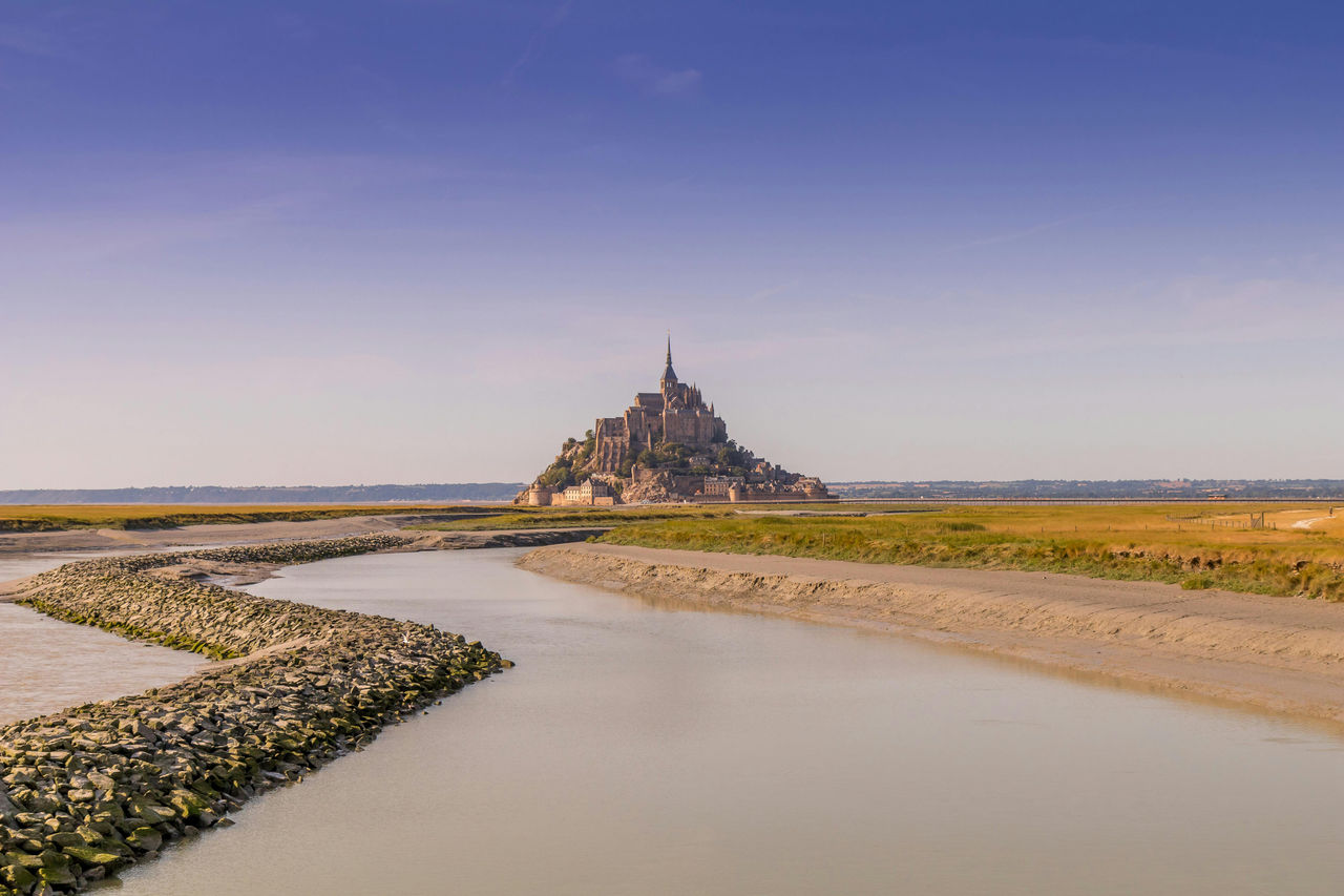 El Mont-Saint-Michel se alza sobre las marismas en la frontera entre Normandía y Bretaña, enmarcado por un río tranquilo y cielos abiertos.