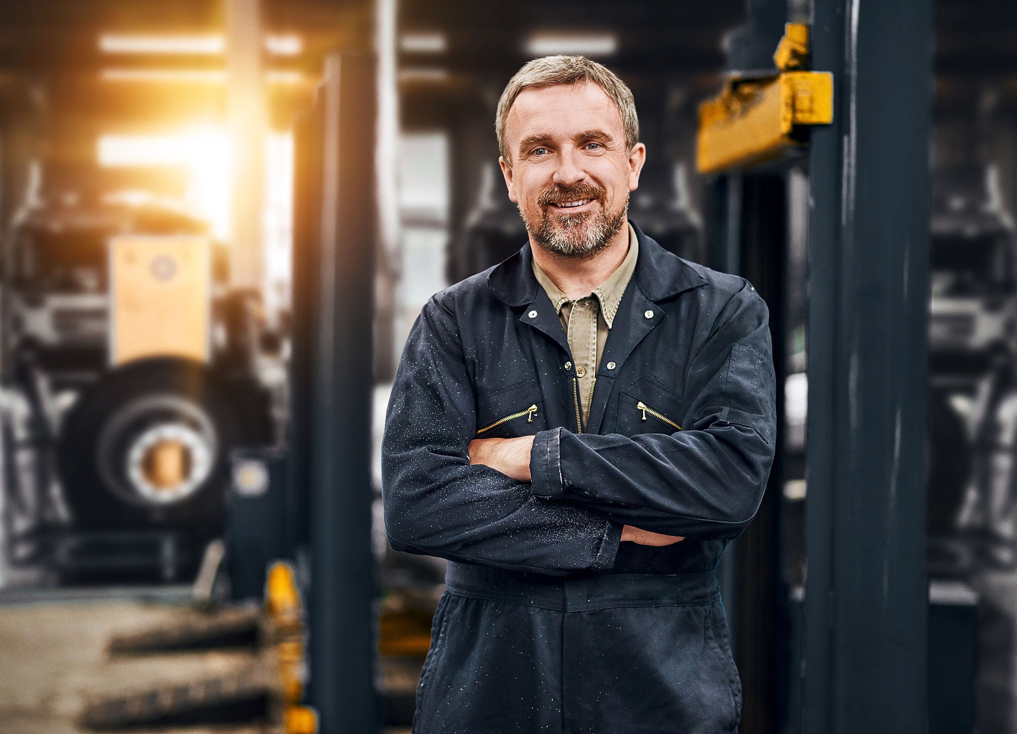 A smiling garage owner stands looking to camera from his busy auto repair shop .