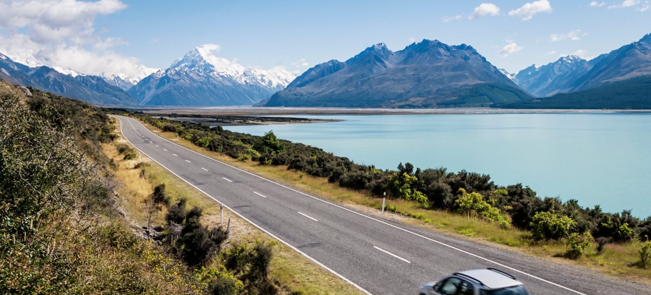 Coche circulando por una carretera con un lago y montañas en el fondo